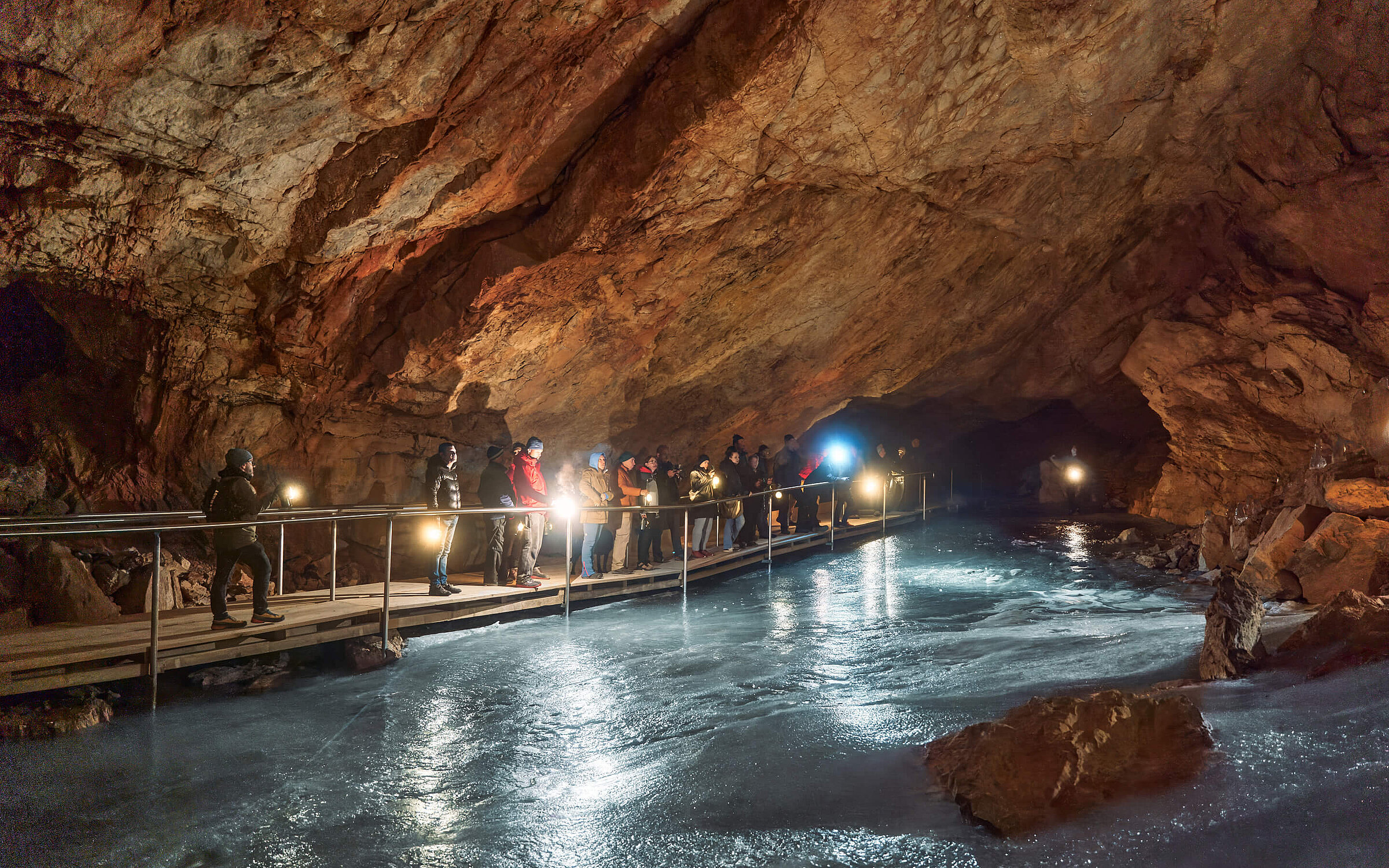 Eine Gruppe von Besuchern geht über eine schmale Holzbrücke in der Eisriesenwelt Werfen. Die große Höhle hat einen eisigen Boden und felsige Wände.