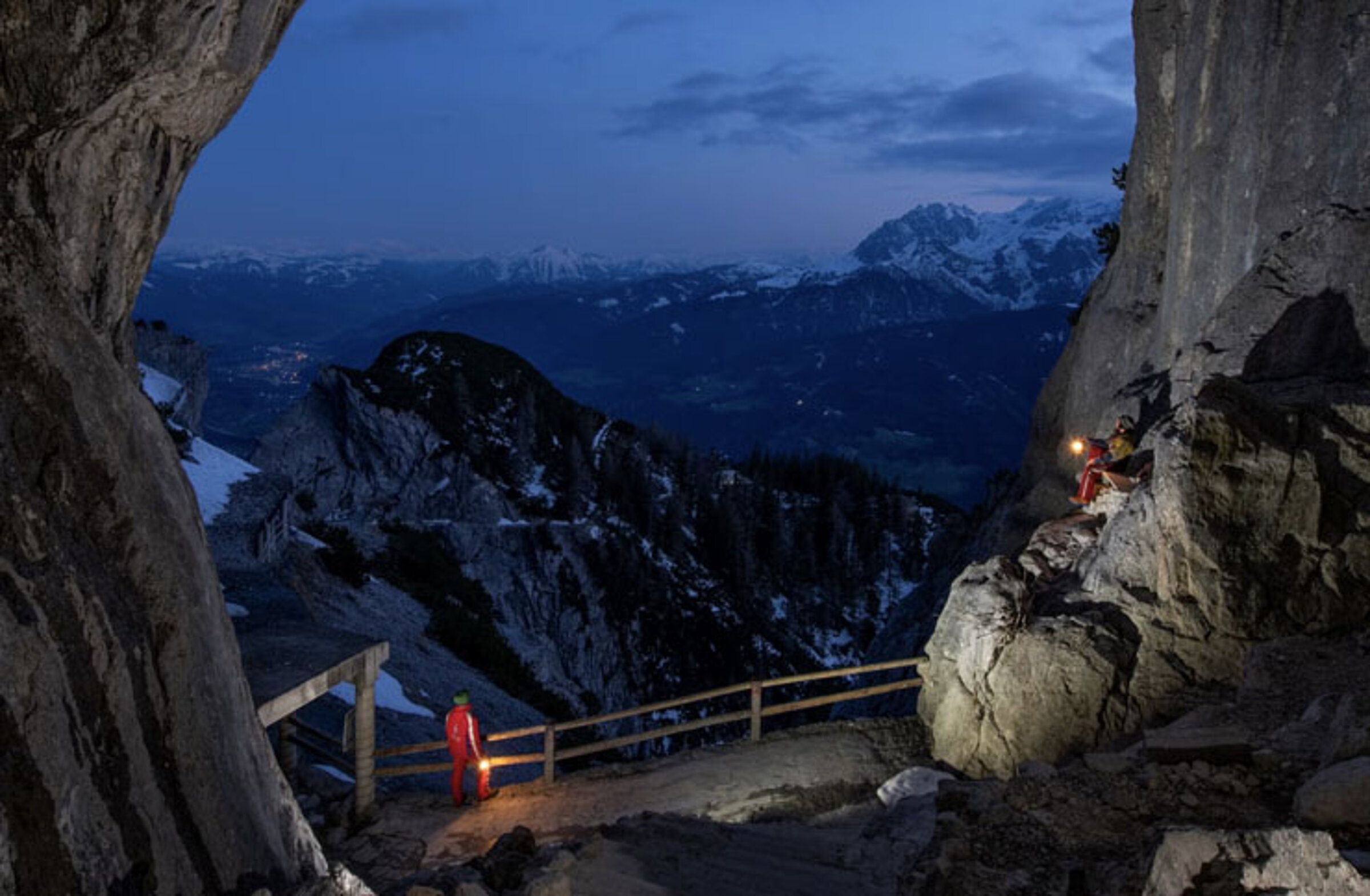 Two mountaineers with lamps stand at dusk at the entrance to the rocky mountain cave to the Eisriesenwelt. A panorama of snow-covered peaks stretches out before them under an atmospheric evening sky.