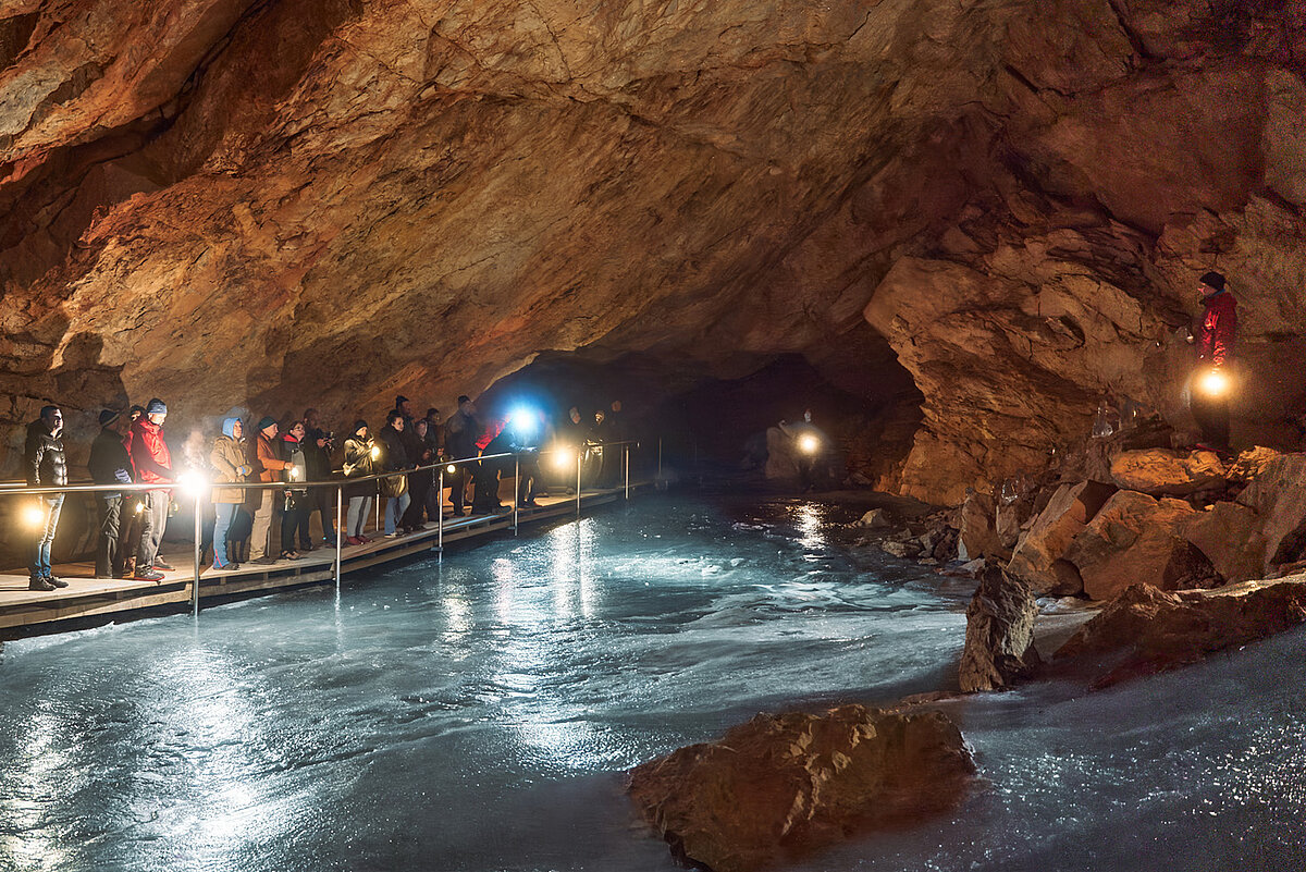  Eine Gruppe von Menschen erkundet die Eishöhle mit großen Eiszapfen und vereisten Wänden. Eine Person steht auf dem Eis und hält eine Laterne in der Hand.