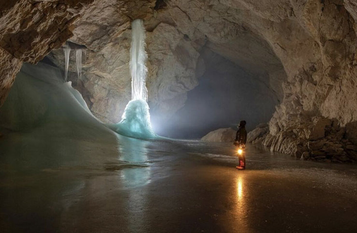 A person with a carbide lamp stands in front of a pillar-like ice formation in the enormous Ice Giant World cave. The smooth, icy cave floor reflects the light in shimmering reflections and enhances the impressive atmosphere.