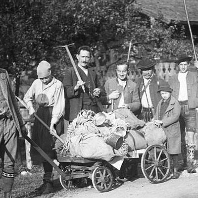 A black and white photo shows a group of people standing outside behind a cart filled with sacks and milk cans. Some are holding agricultural tools. A house with a fence can be seen in the background.
