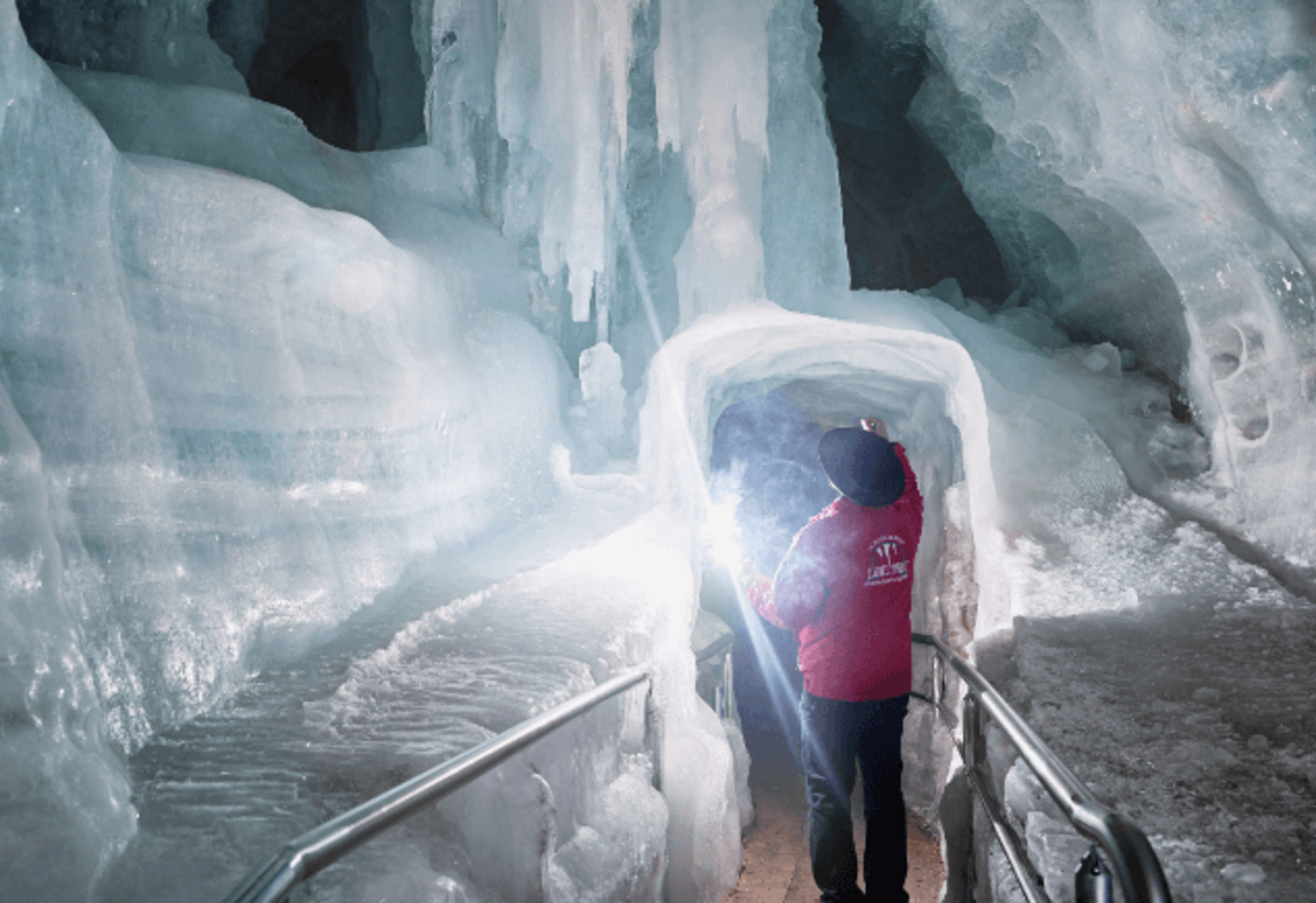 A cave guide in a red jacket walks through an icy tunnel in the Ice Giant World with a carbide lamp in her hand. The walls and ceiling are made of thick ice, icicles hang down and a metal railing lines the path for visitors.