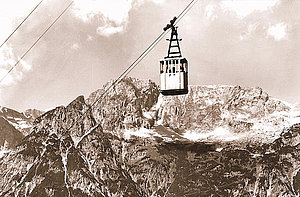 A historic cable car suspended on ropes glides over rugged, snow-covered peaks under a partly cloudy sky.
