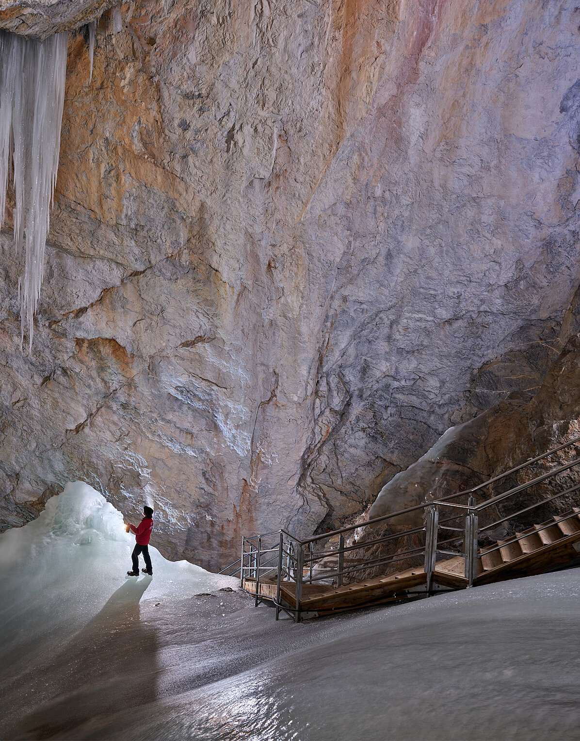 Eine Person in einer roten Jacke steht neben einer Treppe in einer großen Höhle mit Eiswänden und riesigen Eiszapfen, die von der Decke hängen.