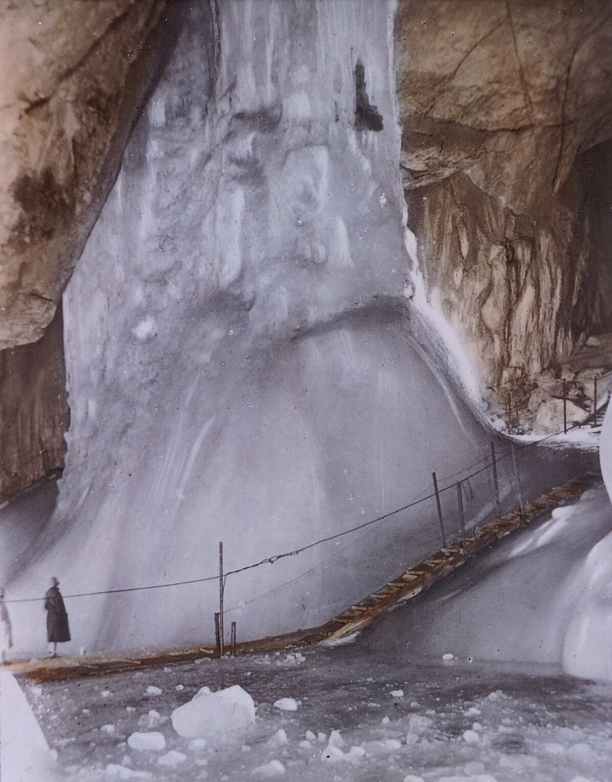Eine Person steht in der Nähe eines gewaltigen Eisformation in der Eisriesenwelt, wo ein Holzsteg und ein Seilgeländer die Besucher durch Eis- und Felswände führen – eine unvergessliche Szene voller Geschichte.