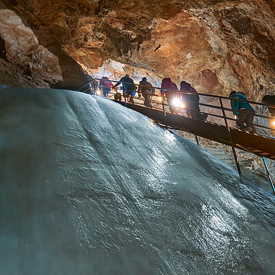 Eine Gruppe von Menschen steigt eine Treppe in der Eisriesenwelt hinauf, neben einem riesigen eisigen Abhang und schroffen braunen Felswänden.