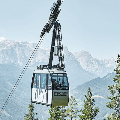 A cable car travels above treetops, with mountains visible in the distance. The cable car is labeled “Eisriesenwelt” and carries several passengers.