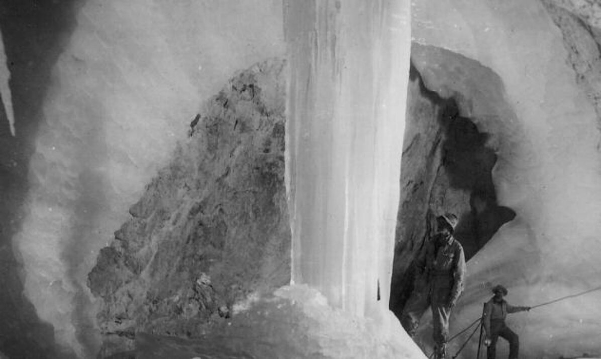 Black and white photo of two people in an ice cave, standing next to an imposing, vertical column of ice. The cave walls and floor are covered in snow and ice, creating an impressive, dramatic icy landscape.