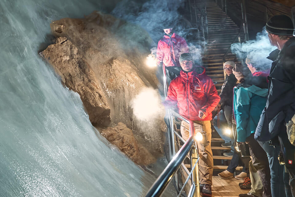 Eine Gruppe von Besuchern in warmer Winterkleidung geht auf einer Holztreppe im Inneren der Eisriesenwelt Werfen hinunter, direkt vorbei an einer gewaltigen, beleuchteten Eiswand.