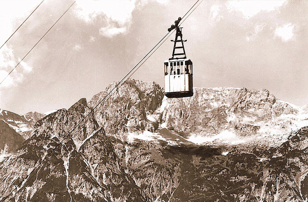 A historic cable car suspended on ropes glides over rugged, snow-covered peaks under a partly cloudy sky.