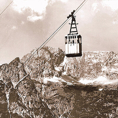 Eine an Seilen aufgehängte historische Seilbahn gleitet unter einem teilweise bewölkten Himmel über schroffe, schneebedeckte Gipfel.