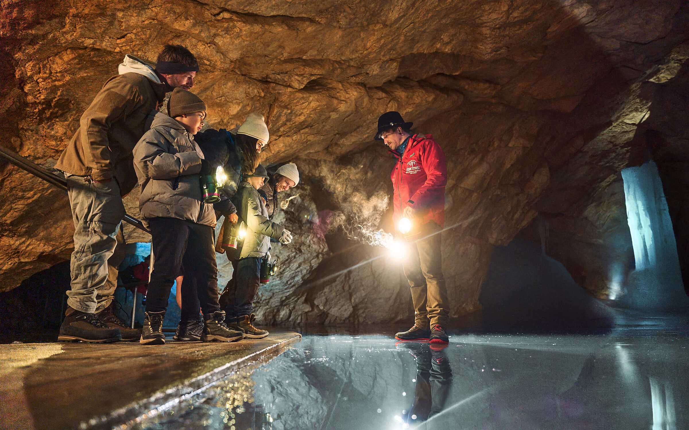 Eine Gruppe von Besuchern, darunter auch Kinder, erkundet die Eisriesenwelt Werfen und betrachtet eine spiegelglatte Eisfläche im Höhleninneren.
