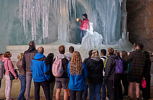 A group of people in jackets stand in the Ice Giant World cave and listen to a cave guide in a red jacket holding a carbide lamp while fascinating guides reveal the history behind the frozen formations.