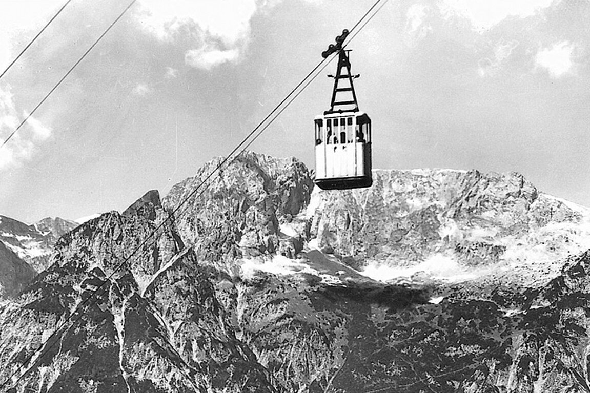 Eine alte Seilbahn, die an Drähten hängt, fährt unter einem teilweise bewölkten Himmel über schroffe, schneebedeckte Berggipfel. 
