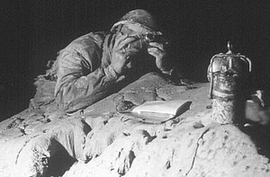 Black and white photo of a speleologist wearing a helmet, lying on a rock in a dimly lit cave. His face is partially covered by a camera. Next to him are a lamp and an open notebook.