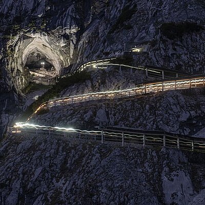 A winding mountain path, illuminated by strips of light at night, leads up to the large cave entrance of the Ice Giant World, which is impressively situated between dramatic rocky cliffs.