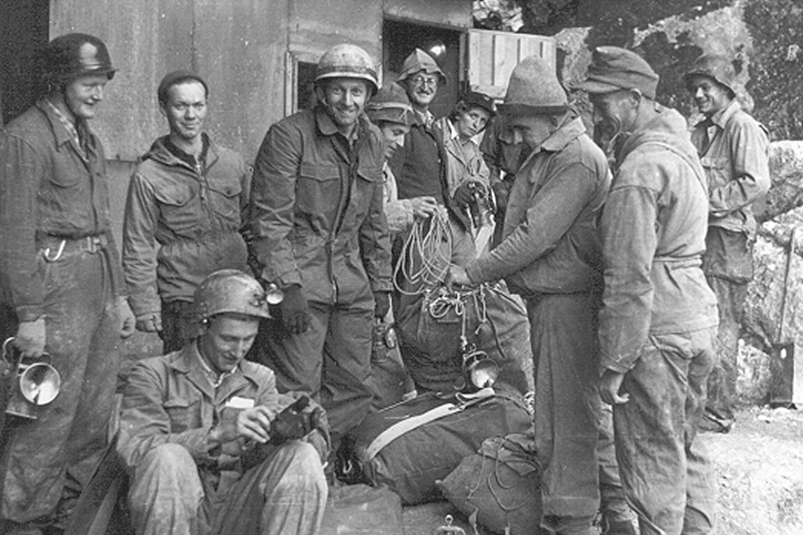Eine Schwarz-Weiß-Aufnahme einer Gruppe von Menschen, die sich in der Nähe einer Hütte versammelt haben, lächeln und Kletterausrüstung in den Händen halten.