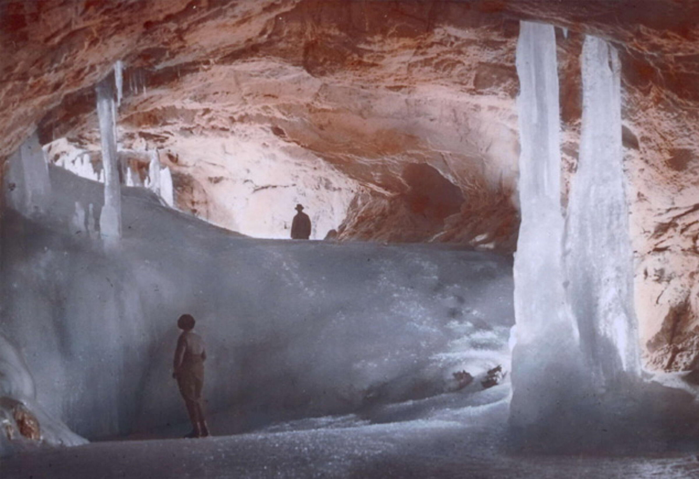 Zwei Menschen stehen in der Eisriesenwelt, einer riesigen Höhle mit eisigem Boden und hoch aufragenden Eisformationen. Die rotbraunen Wände der Höhle bilden einen Kontrast zum Eis und laden Besucher ein, ihre Geschichte bei spannenden Führungen zu erkunden.
