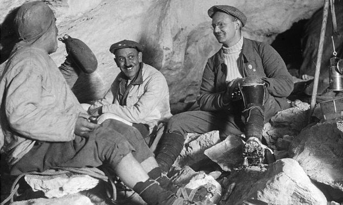 Black and white photo of three men in early 20th century clothing sitting smiling on rocks in a cave and drinking from bottles.