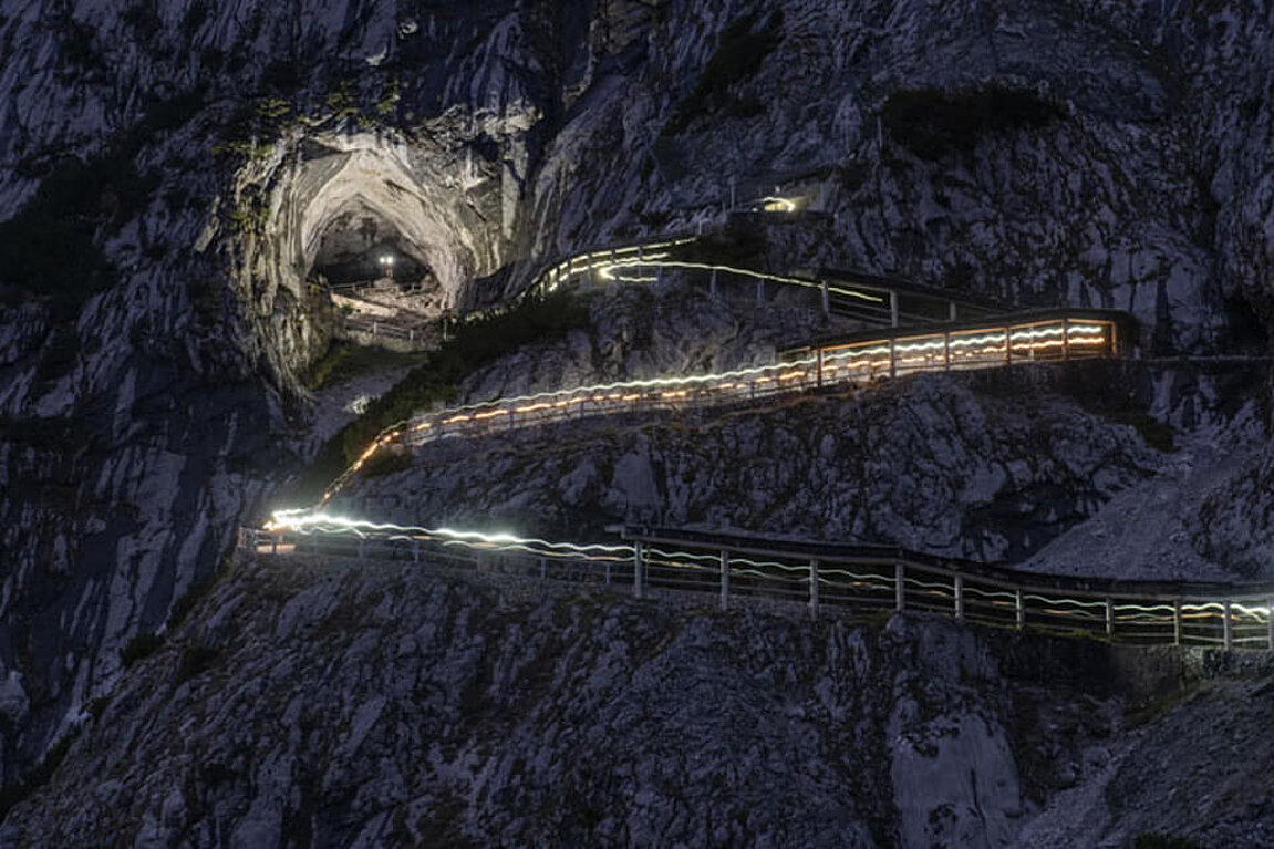 Eine Nachtaufnahme zeigt den beleuchteten Weg und den imposanten Eingang zur Eisriesenwelt in Werfen, der sich hell gegen die dunkle Berglandschaft abhebt.
