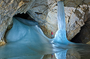A cave guide in a red jacket stands in the cave and looks up at a large frozen ice formation surrounded by ice and rock faces with reflections on the icy floor.