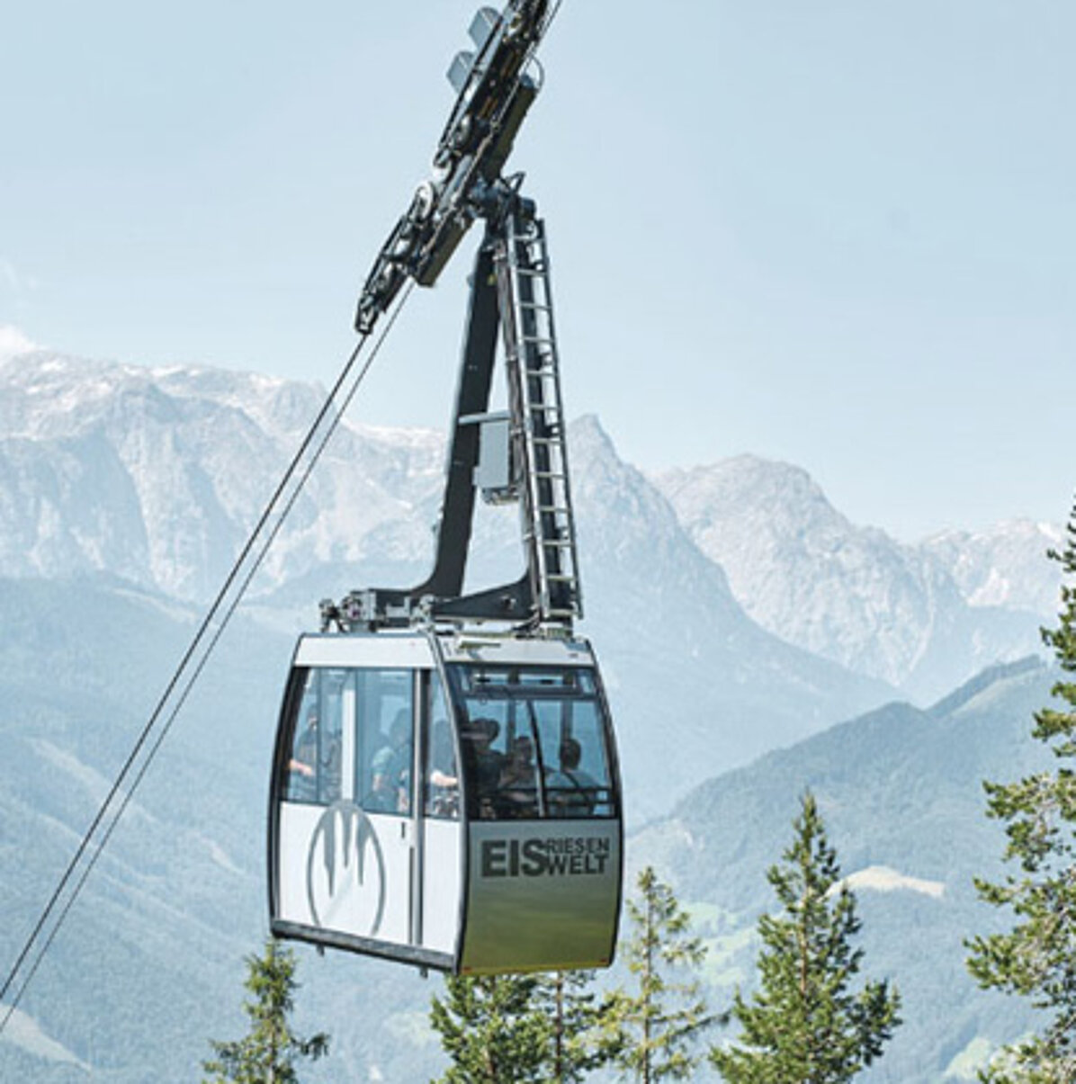 Eine Seilbahn fährt über Baumwipfel, im Hintergrund sind Berge in der Ferne zu sehen. Die Seilbahn trägt die Aufschrift „Eisriesenwelt“ und befördert mehrere Passagiere.