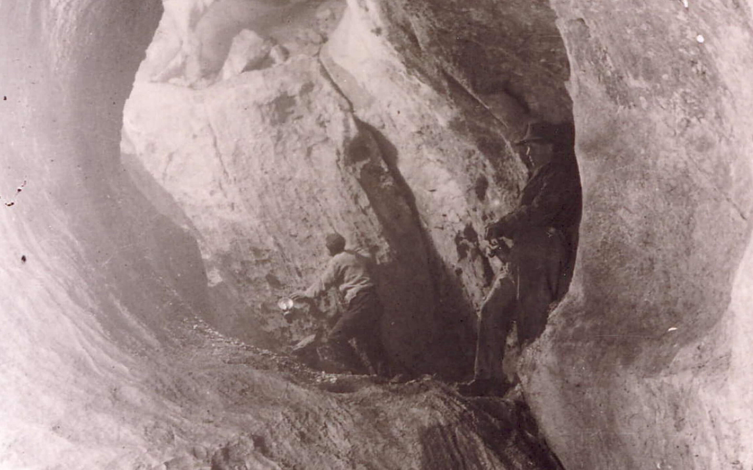 Historische Schwarz-Weiß-Aufnahme aus der Eisriesenwelt Werfen: Zwei Personen in einer großen, glatten Felsformation – eine geht tiefer in die Höhle hinein, während die andere mit Hut an der rechten Felswand lehnt.