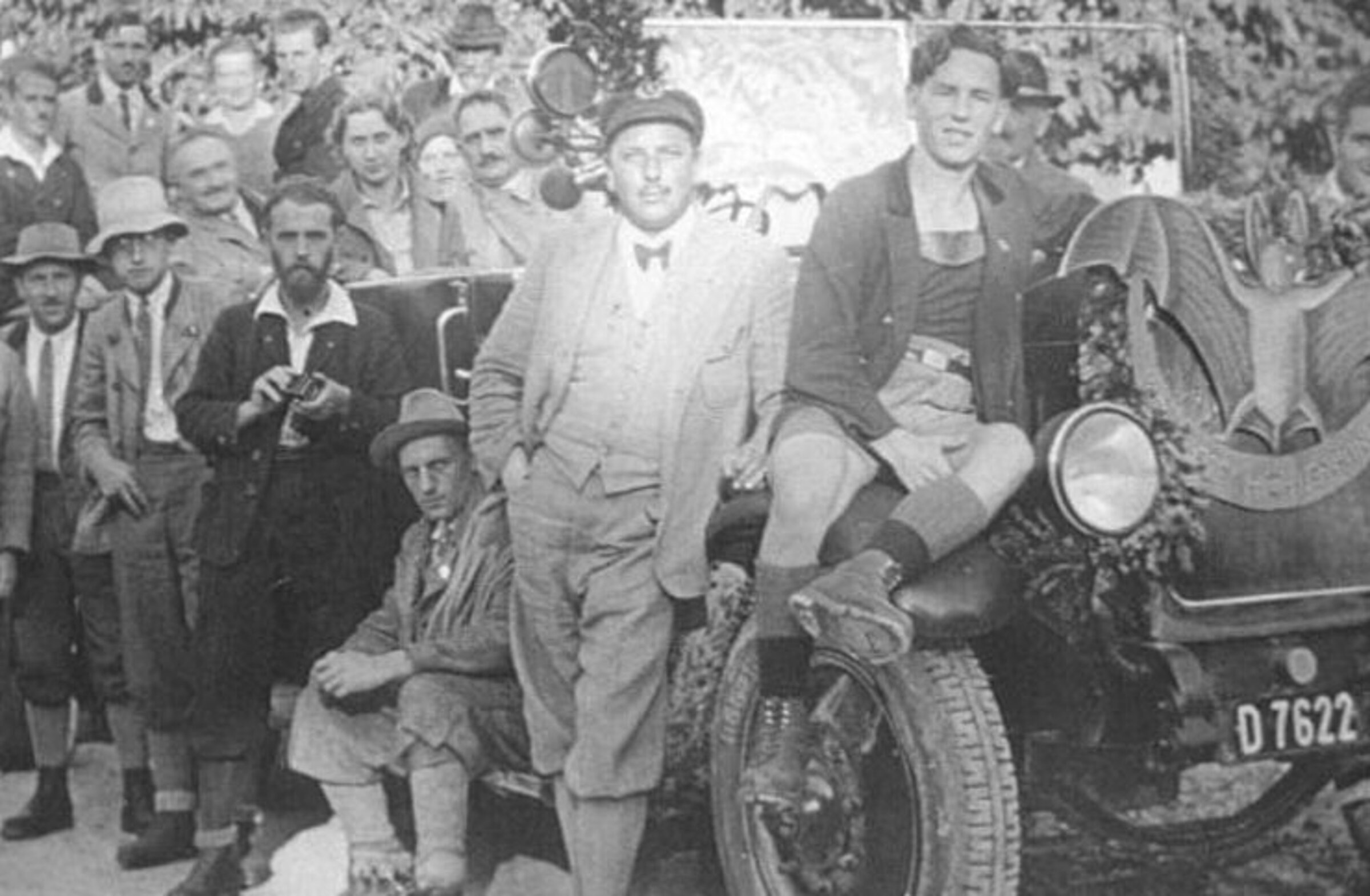 Black and white photo from the early 20th century: A group of people, mostly men, pose around a festively decorated car. Two men are sitting on the car, while others standing or sitting nearby are smiling and looking at the camera.