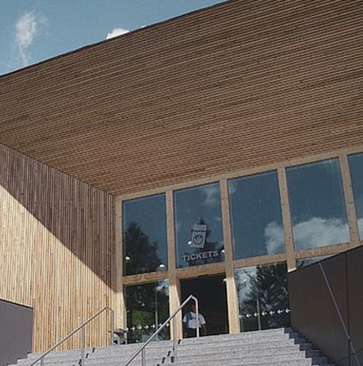 A modern visitor centre with vertical wooden slats, large glass windows and a ticket sales sign above the entrance welcomes visitors to the Eisriesenwelt cable car. On a sunny day, stone steps lead up to the door.
