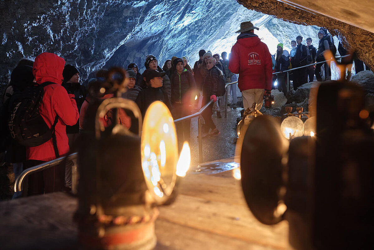 Eine Gruppe von Menschen erkundet, angeführt von einem Führer in roter Jacke und Hut, die Eisriesenwelt. Im Vordergrund stehen alte Karbidlampen.