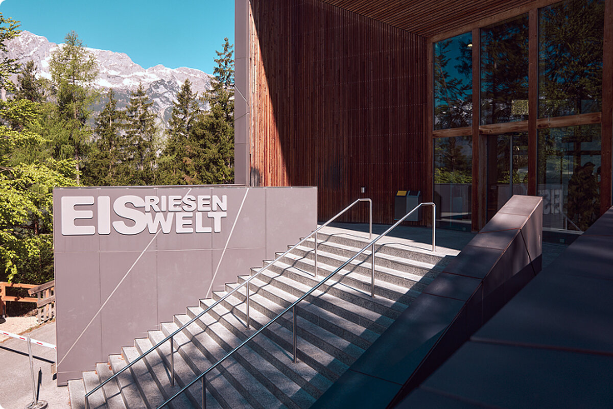 Aussenansicht des Besucherzentrums mit Treppe zum Eingang. Auf einem Schild neben der Treppe steht „EISRIESENWELT“. 