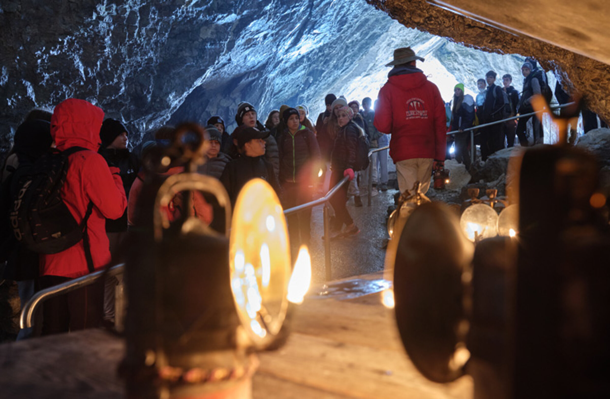 Eine Gruppe von Menschen erkundet, angeführt von einem Führer in roter Jacke und Hut, die Eisriesenwelt. Im Vordergrund stehen alte Karbidlampen.