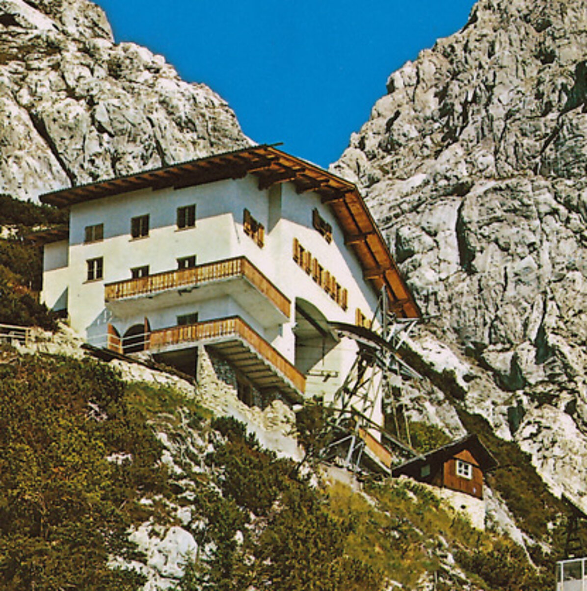 Das Bergstationsgebäude, ein weißes Alpenhaus mit Holzbalkonen, steht an einem Felshang in der Nähe der Eisriesenwelt, eingerahmt von Gebüsch und steilen grauen Felsen unter einem klaren blauen Himmel.