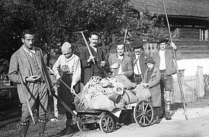A black and white photo shows a group of people standing outside behind a cart filled with sacks and milk cans. Some are holding agricultural tools. A house with a fence can be seen in the background.