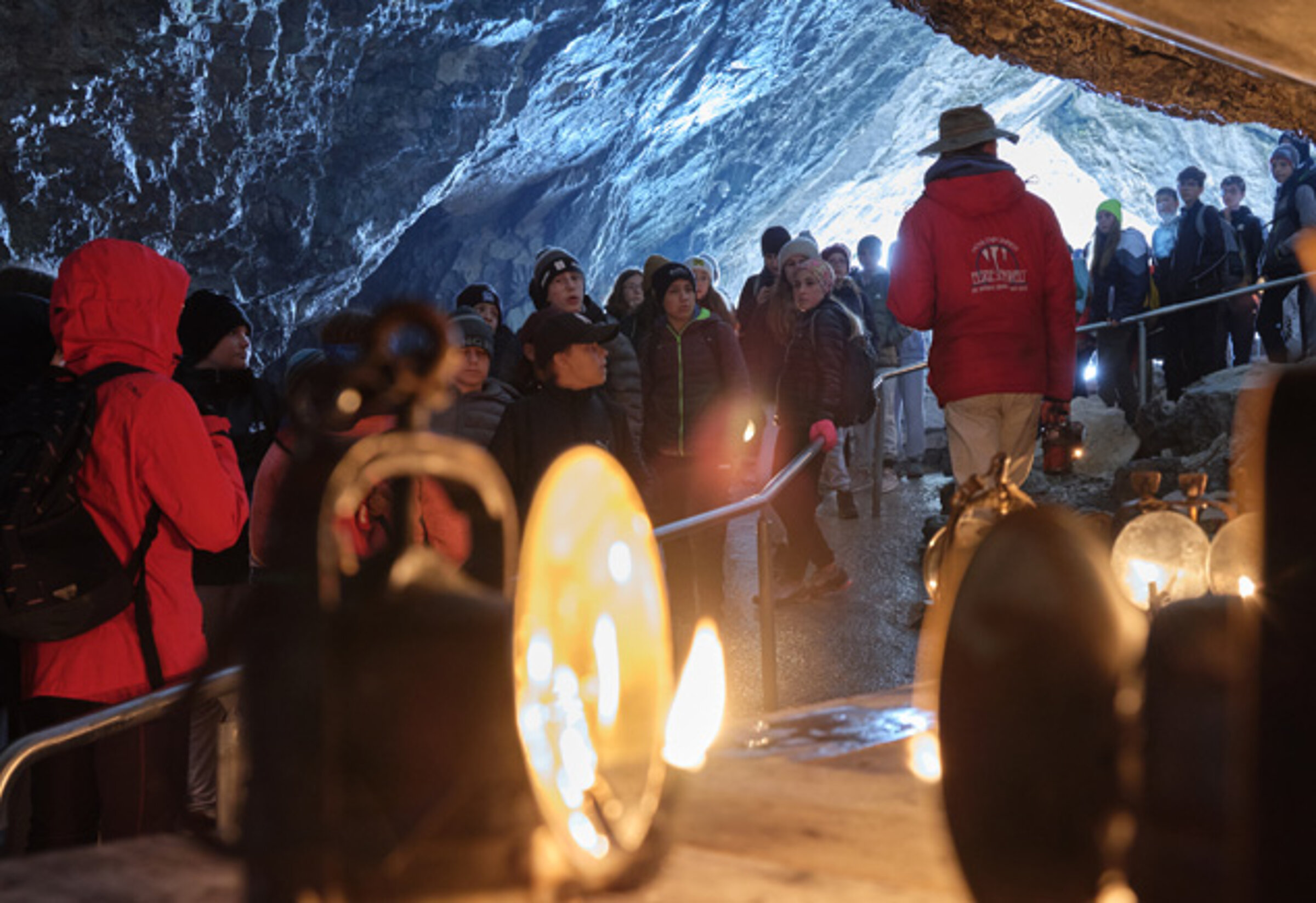 Eine Gruppe von Menschen erkundet, angeführt von einem Führer in roter Jacke und Hut, die Eisriesenwelt. Im Vordergrund stehen alte Karbidlampen.