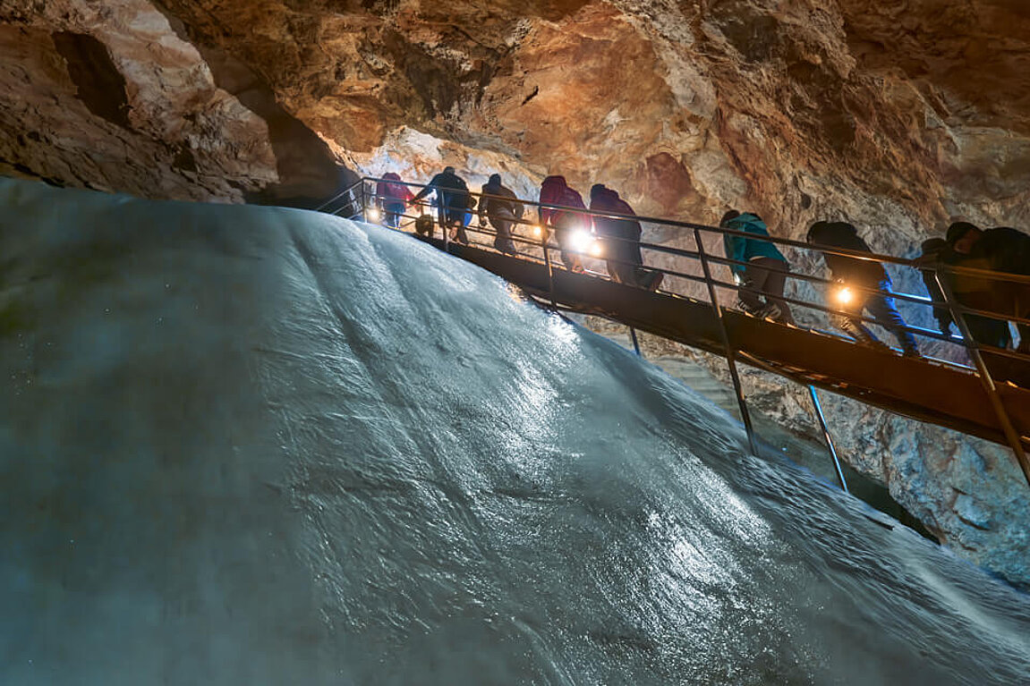 Eine Gruppe von Besuchern in warmer Winterkleidung steigt in der Eisriesenwelt Werfen eine Holztreppe hinauf, die direkt an einer gewaltigen, glatten Eiswand vorbeiführt.