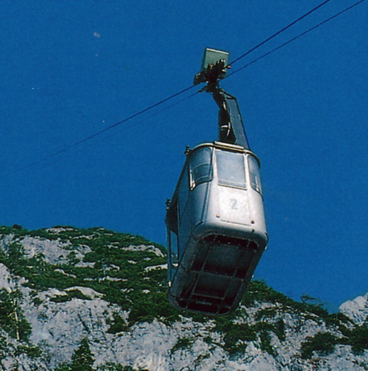Eine silberne Seilbahn mit der Nummer 2 schwebt an einem Kabel vor strahlend blauem Himmel und bewegt sich in Richtung der Berge. Dort befinden sich die berühmte Eisriesenwelt und das Dr.-Friedrich-Oedl-Schutzhaus, eingebettet in felsige und grüne Hänge.