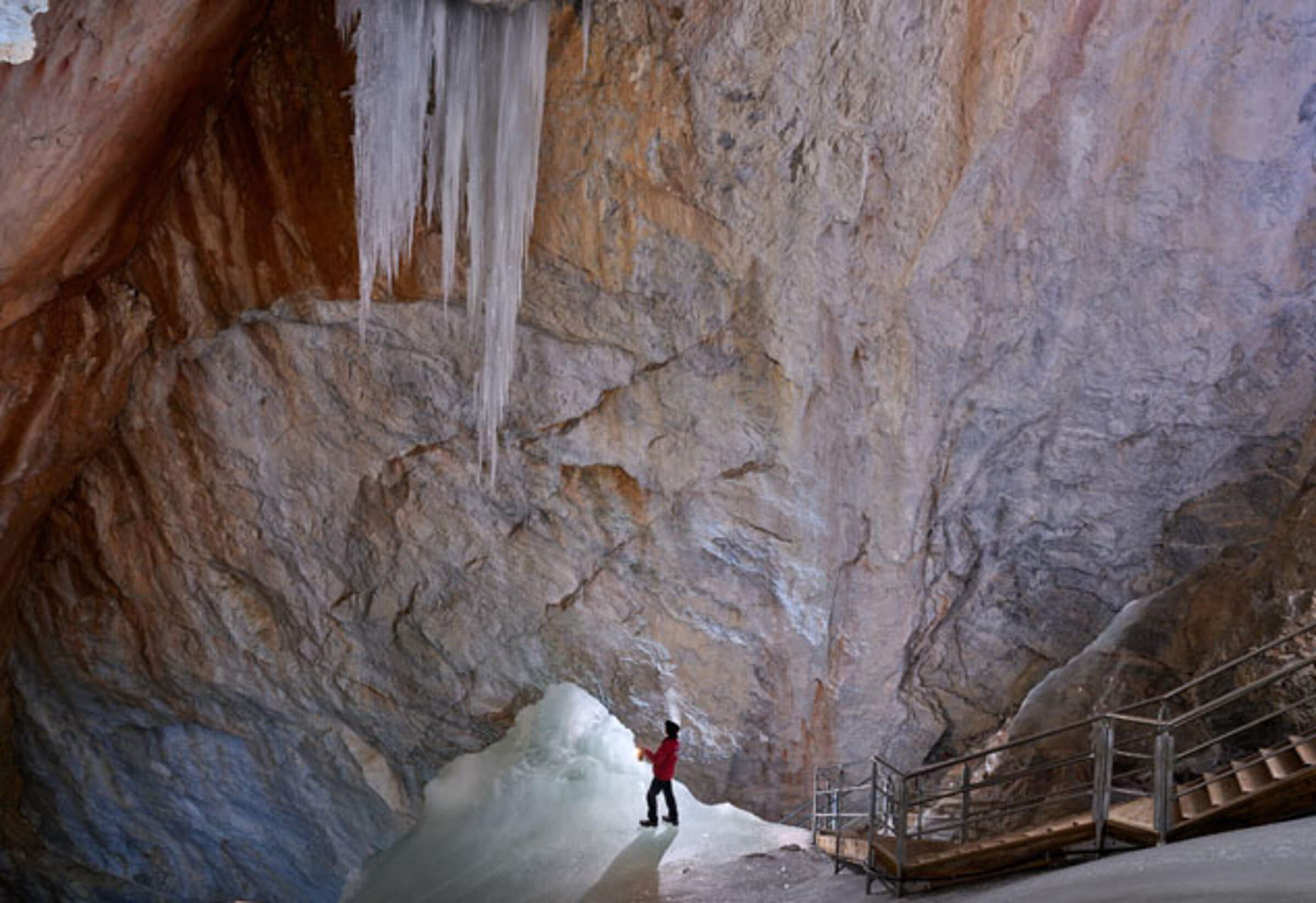 A person in a red jacket stands next to a staircase in a large cave with ice walls and huge icicles hanging from the ceiling.