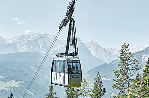 A cable car travels above treetops, with mountains visible in the distance. The cable car is labeled “Eisriesenwelt” and carries several passengers.