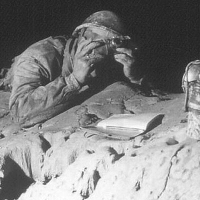 Black and white photo of a speleologist wearing a helmet, lying on a rock in a dimly lit cave. His face is partially covered by a camera. Next to him are a lamp and an open notebook.