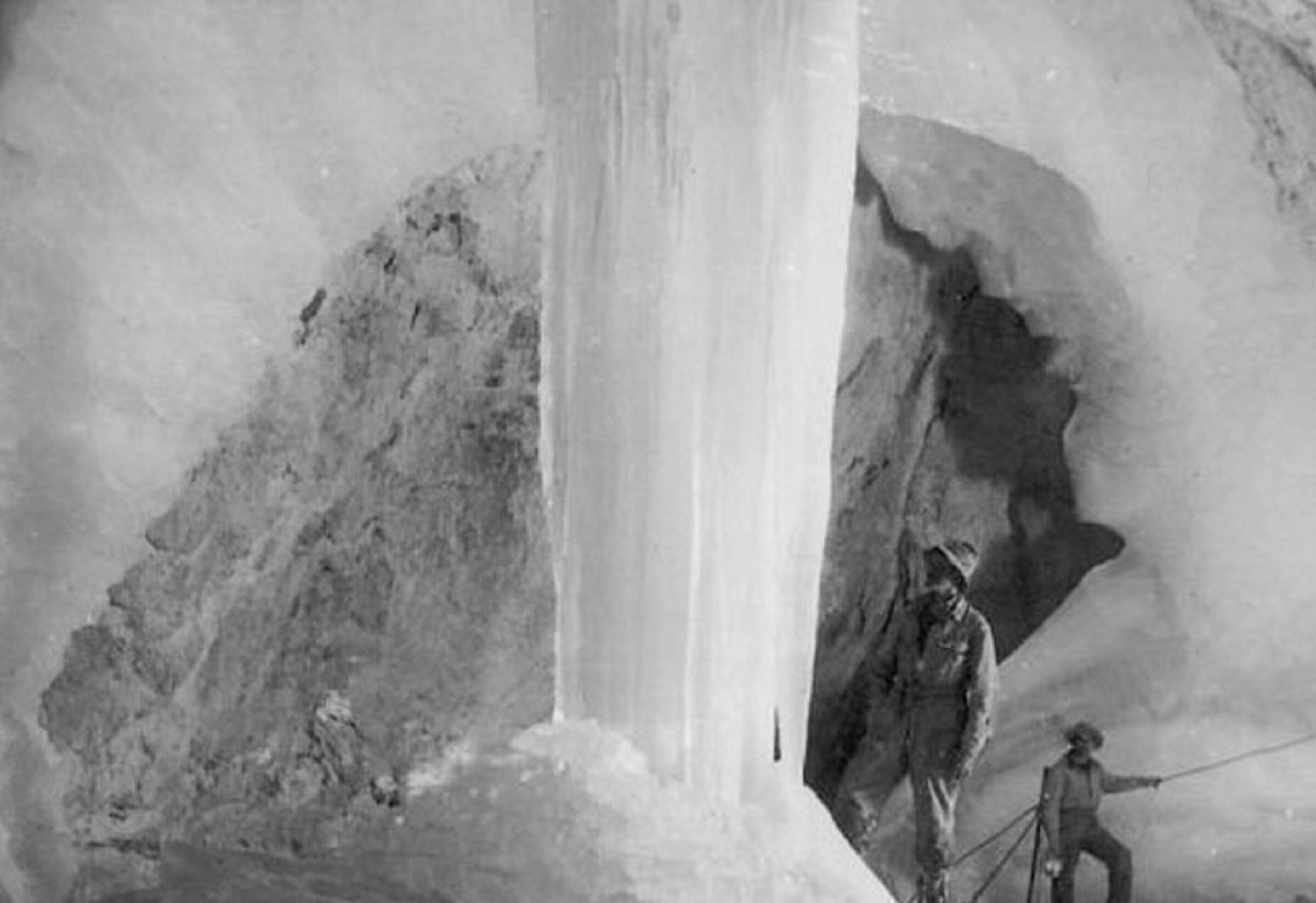 Black and white photo of two people in an ice cave, standing next to an imposing, vertical column of ice. The cave walls and floor are covered in snow and ice, creating an impressive, dramatic icy landscape.